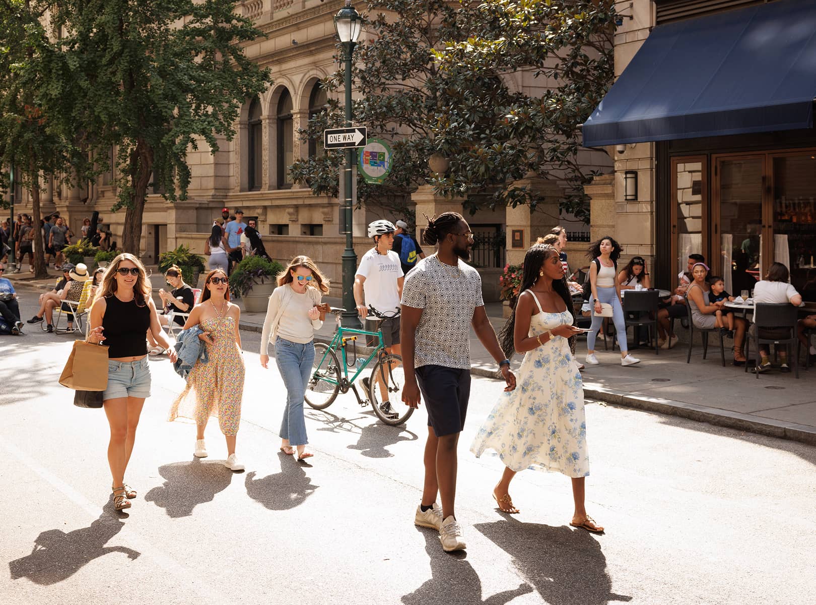 People walk on a sunny street near an outdoor café. The group is casually dressed and smiling, with a cyclist and onlookers in the background. Vibrant, lively atmosphere.