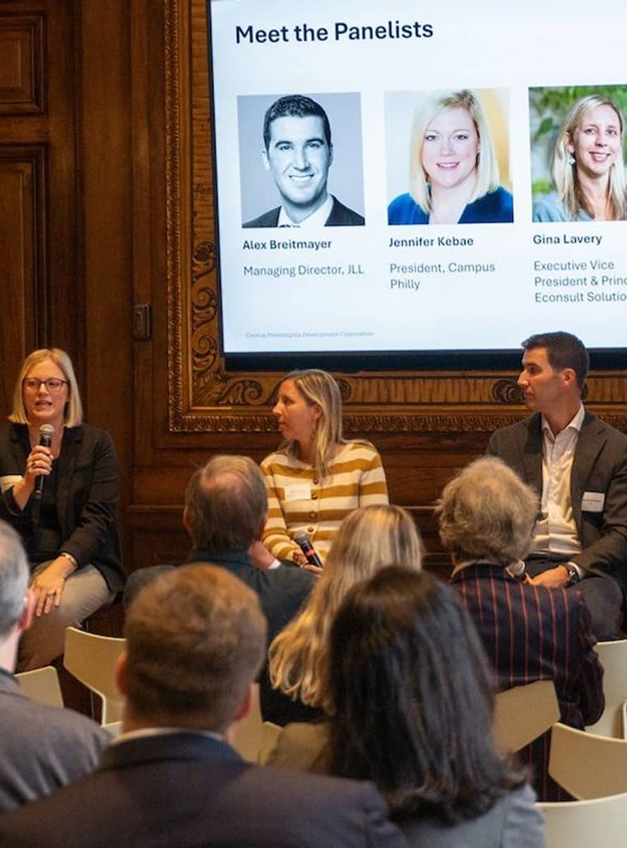 Panelists sit on chairs, facing an audience. A woman speaks into a microphone. A screen behind them displays three panelist photos and names. The setting is formal.