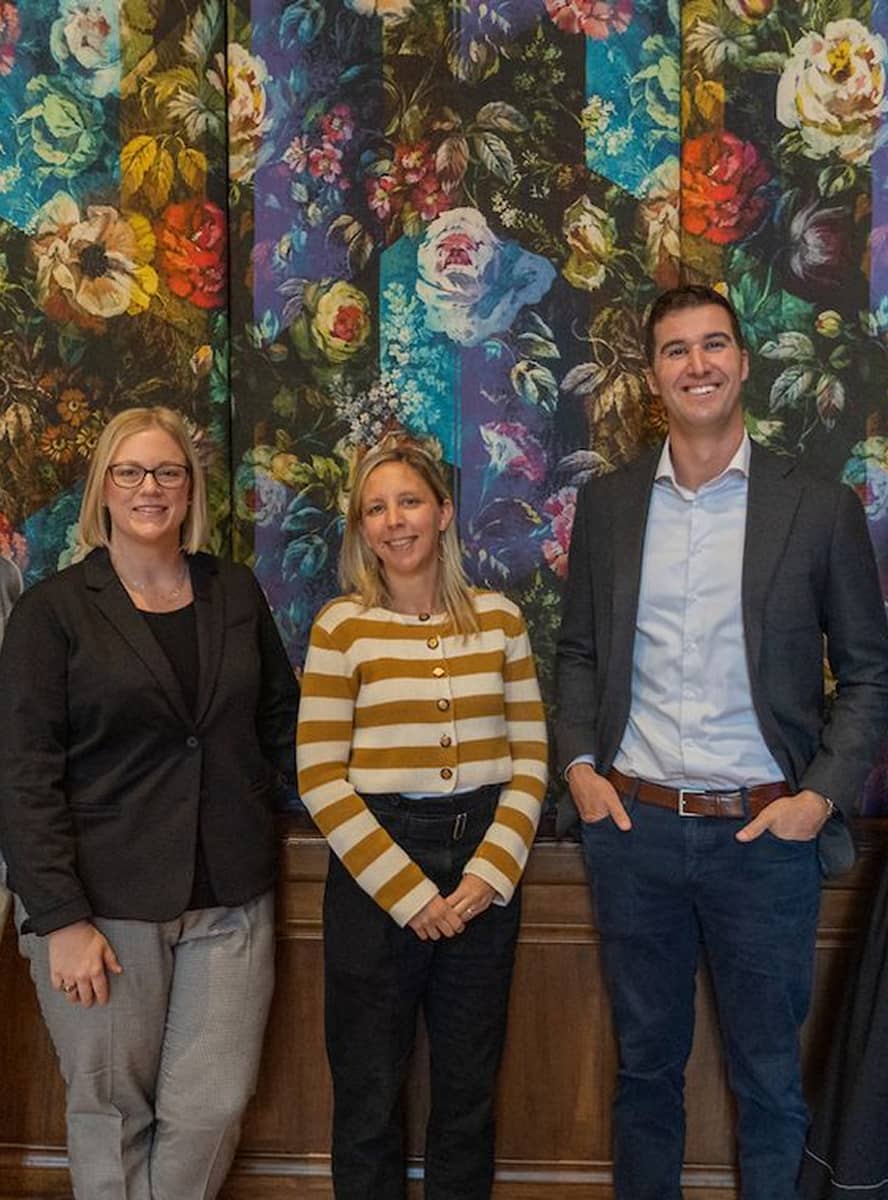 Three people stand smiling in front of a vibrant floral mural. The group includes two women and one man, all dressed in business casual attire.