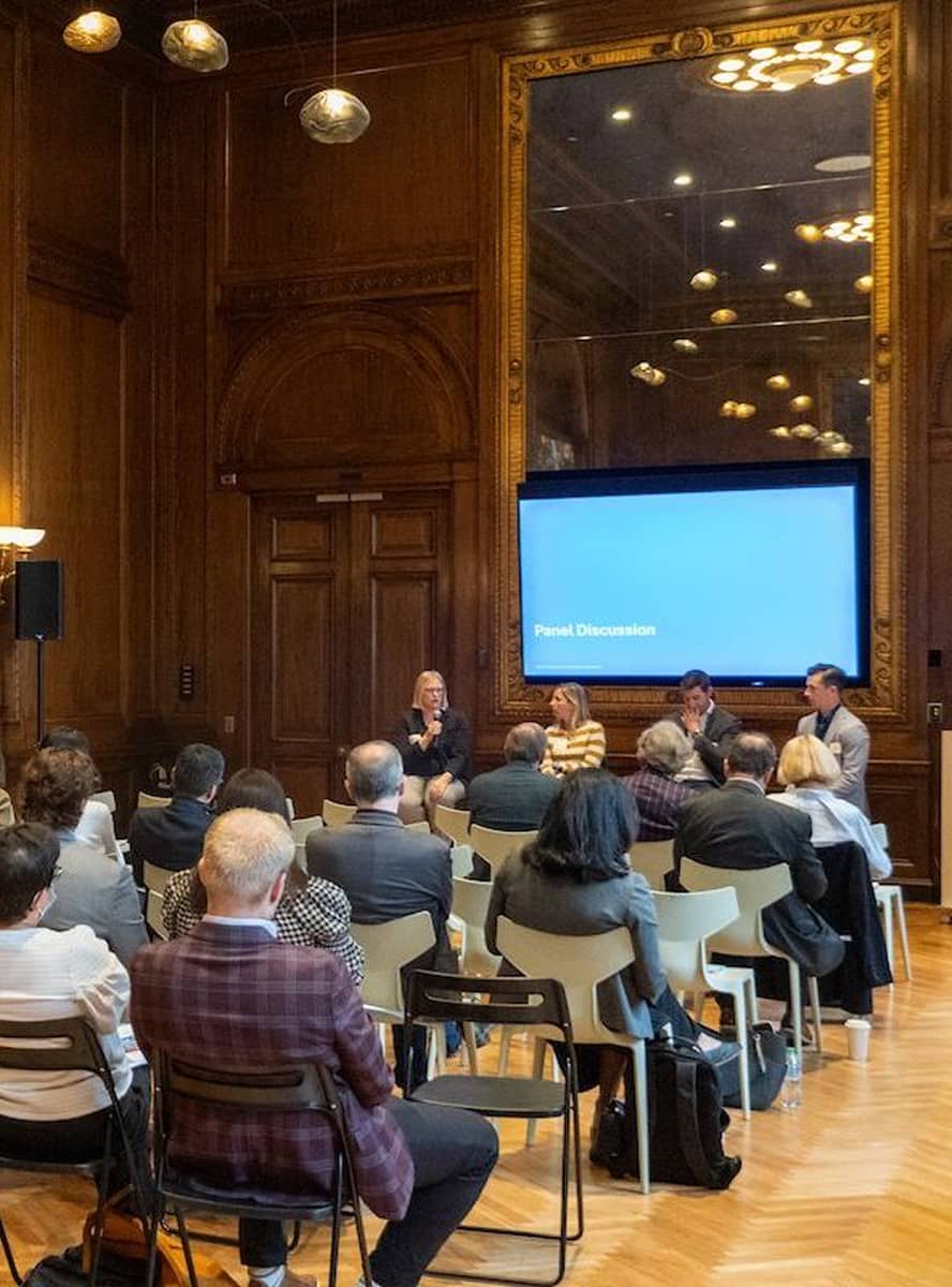 Audience seated in an elegant wood-paneled room with floral artwork, facing a panel discussion on stage. A speaker addresses the group, conveying a professional tone.