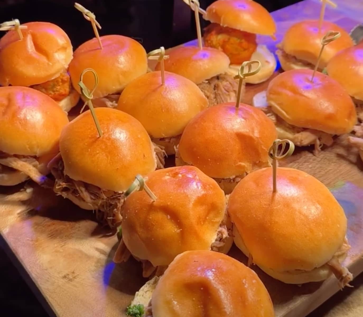 A wooden tray displays a dozen small sandwiches with glossy buns, some filled with pulled meat, others with a fried item, each secured with a skewer.