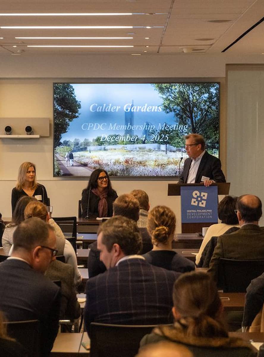 Conference meeting with attendees seated, three speakers at a panel. A large screen displays 