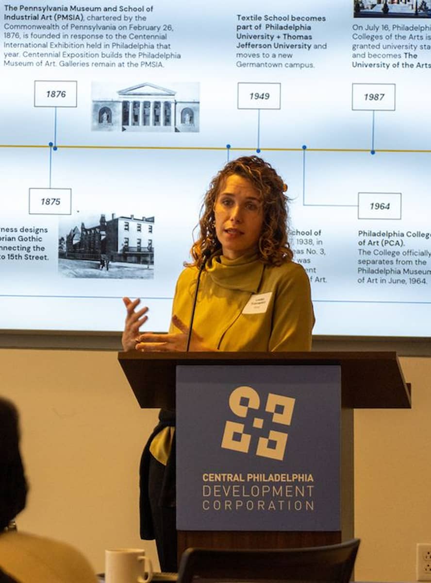 A woman stands at a podium with the Central Philadelphia Development Corporation logo. She is presenting in front of a timeline display, appearing focused.