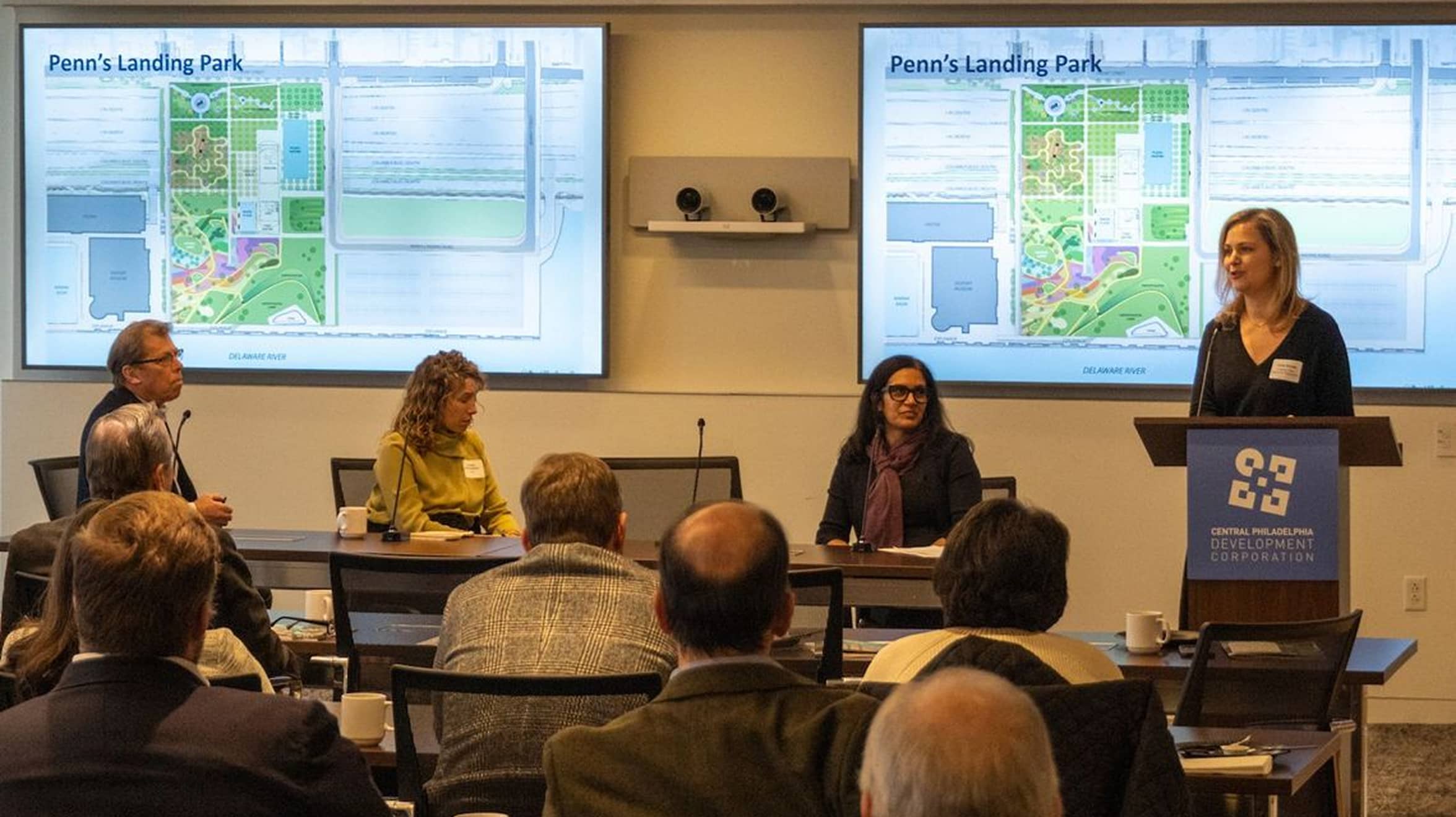A woman stands at a podium speaking to an audience in a conference room. Behind her, two screens display a map titled 