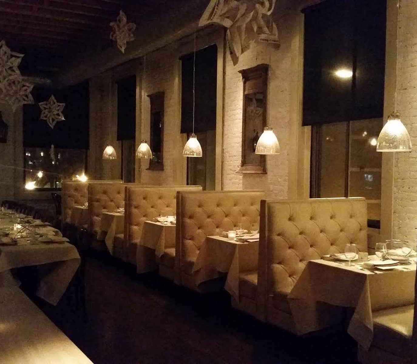 Cozy restaurant interior with plush, beige booth seating and hanging pendant lights. Tables are neatly set, and large windows add warmth and charm.