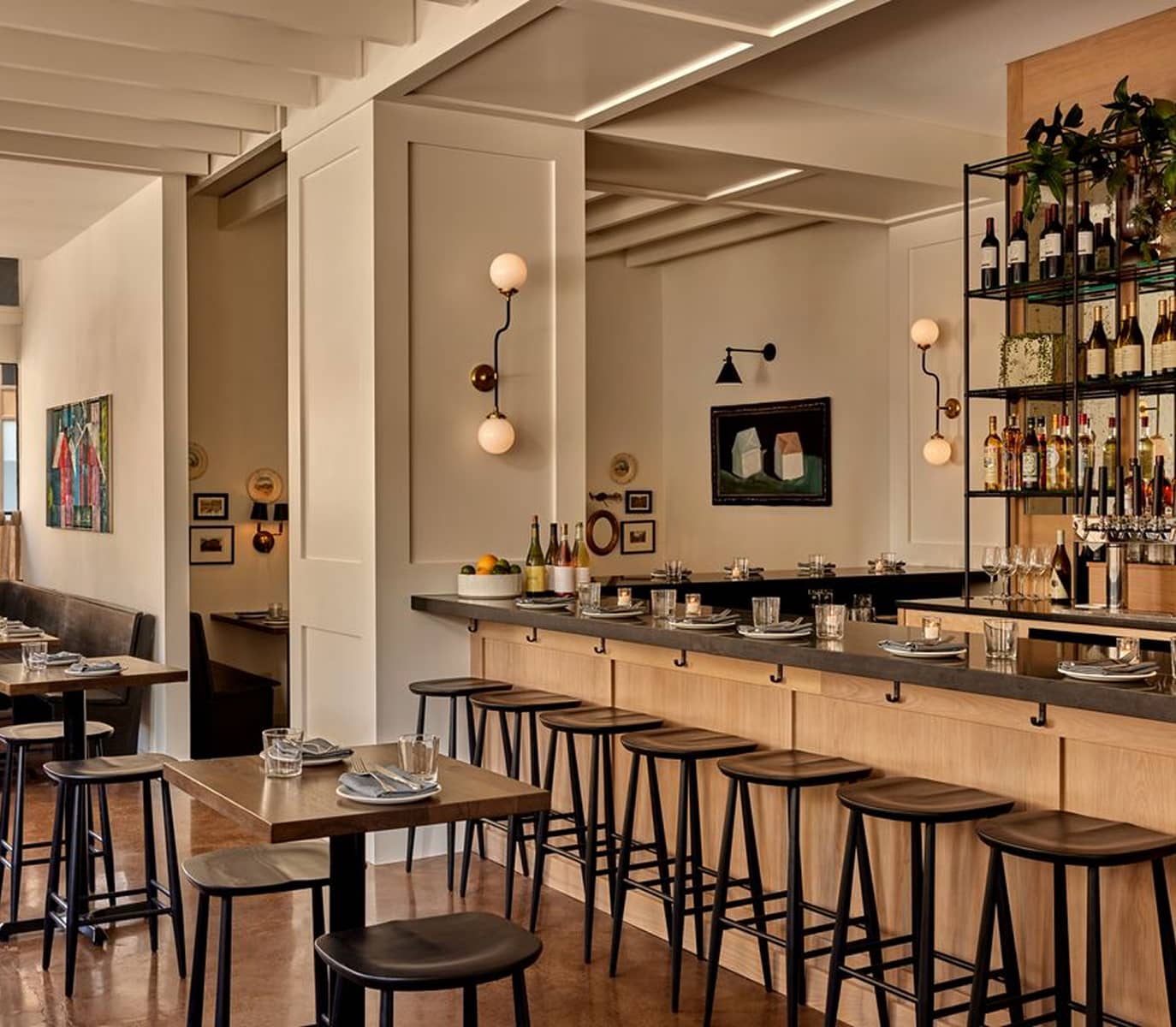 Modern restaurant interior with warm lighting, featuring a sleek bar with high stools, art on walls, and tables set with glasses and plates, creating a cozy ambiance.