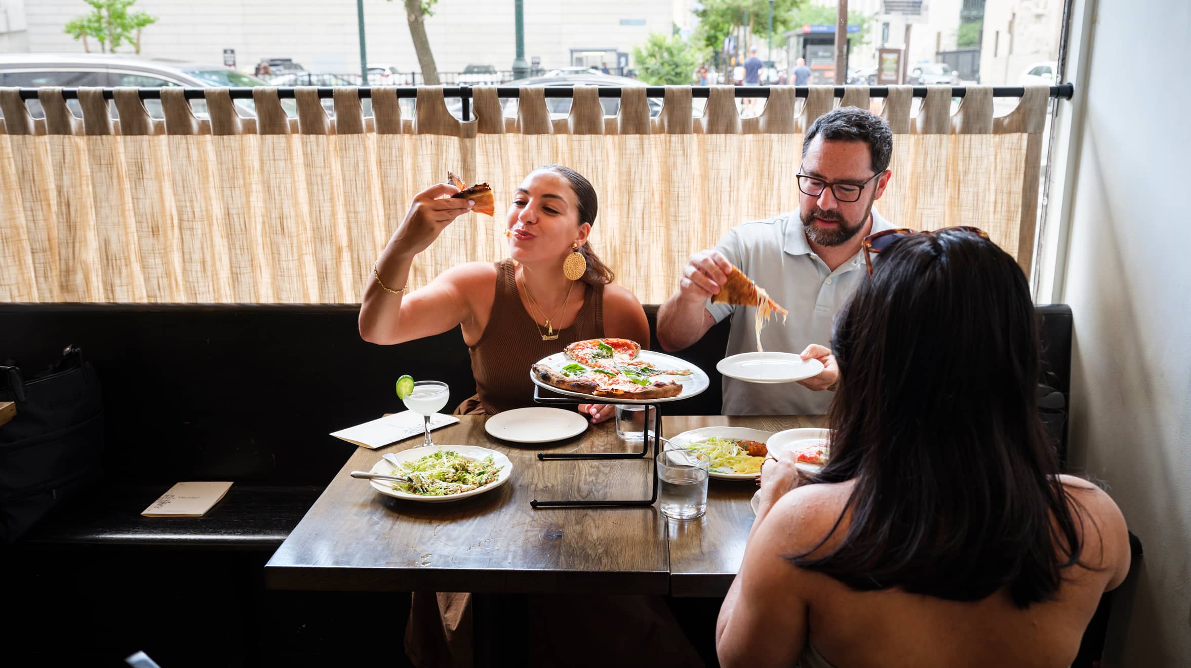 Three people enjoying a meal at a restaurant. A woman on the left eats pizza, a man serves himself, and a woman with her back to the camera looks on.
