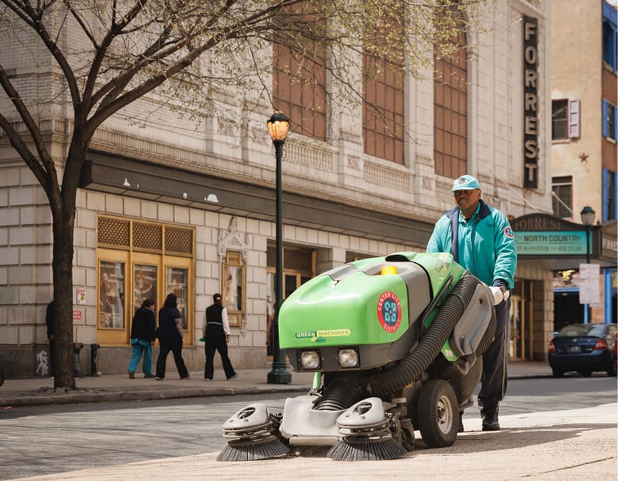A street cleaner operates a green machine, sweeping a city sidewalk. Nearby buildings and a theater marquee are visible, with pedestrians walking by.
