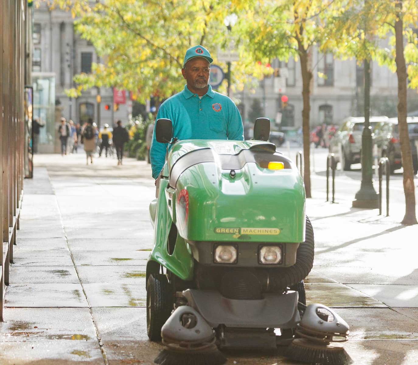 A man in a blue uniform operates a green street cleaning machine on a sunlit city sidewalk. Trees line the street, and pedestrians walk in the background.