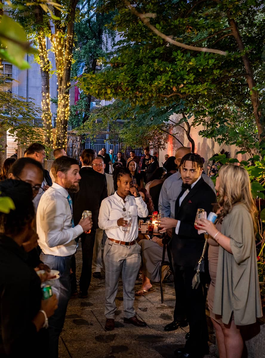 Outdoor evening gathering with people in formal attire under trees adorned with string lights. Attendees are socializing, creating a lively, warm atmosphere.
