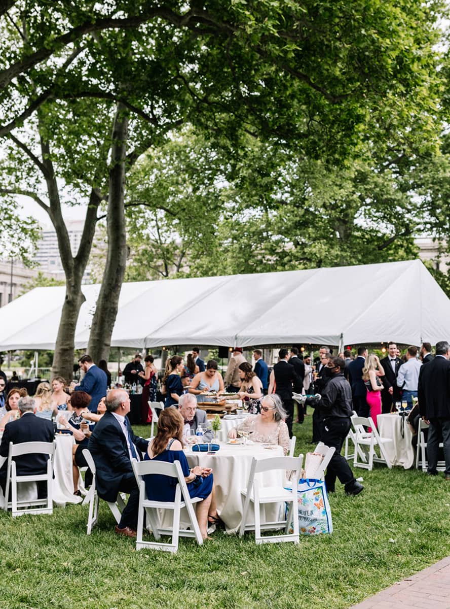 Outdoor gathering with people seated at round tables on a grassy lawn, beneath a large white tent. The scene is lively and social, surrounded by green trees.
