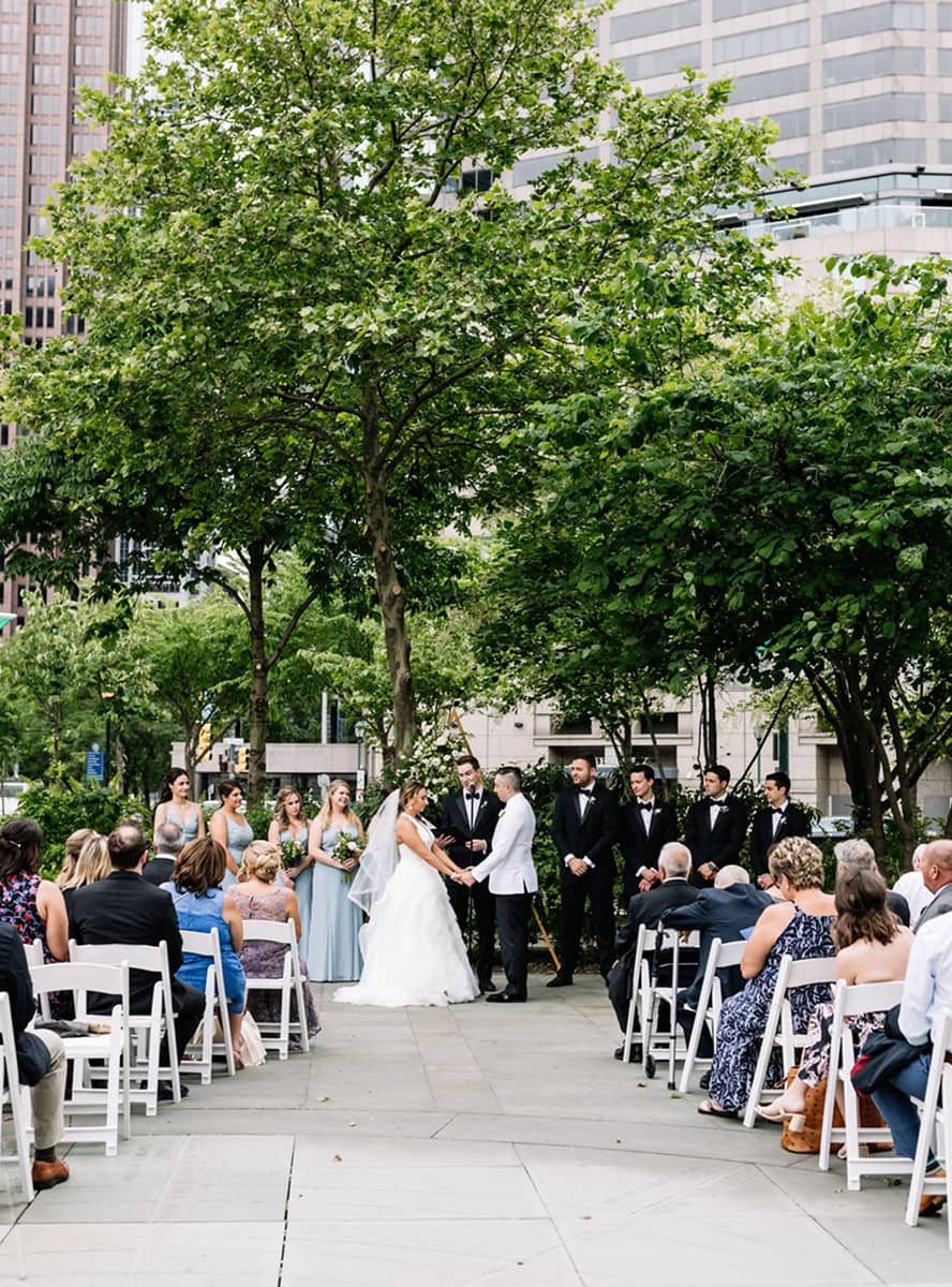 Outdoor wedding ceremony in a park, surrounded by tall trees. The bride and groom stand exchanging vows, with bridesmaids and groomsmen nearby. Guests sit on white chairs.