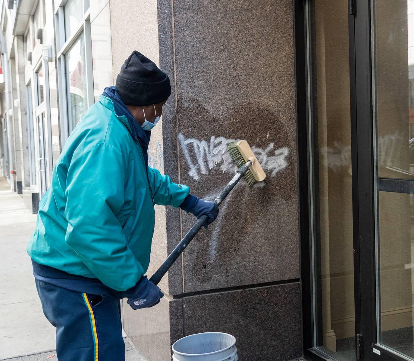 A person in a teal jacket and black beanie is scrubbing graffiti off a brown stone wall with a brush. They hold a bucket nearby, focused on cleaning.