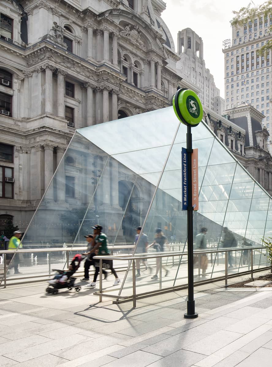 A glass triangular entrance to a subway is set against a historic building. People pass by, including a person pushing a stroller. The mood is lively.