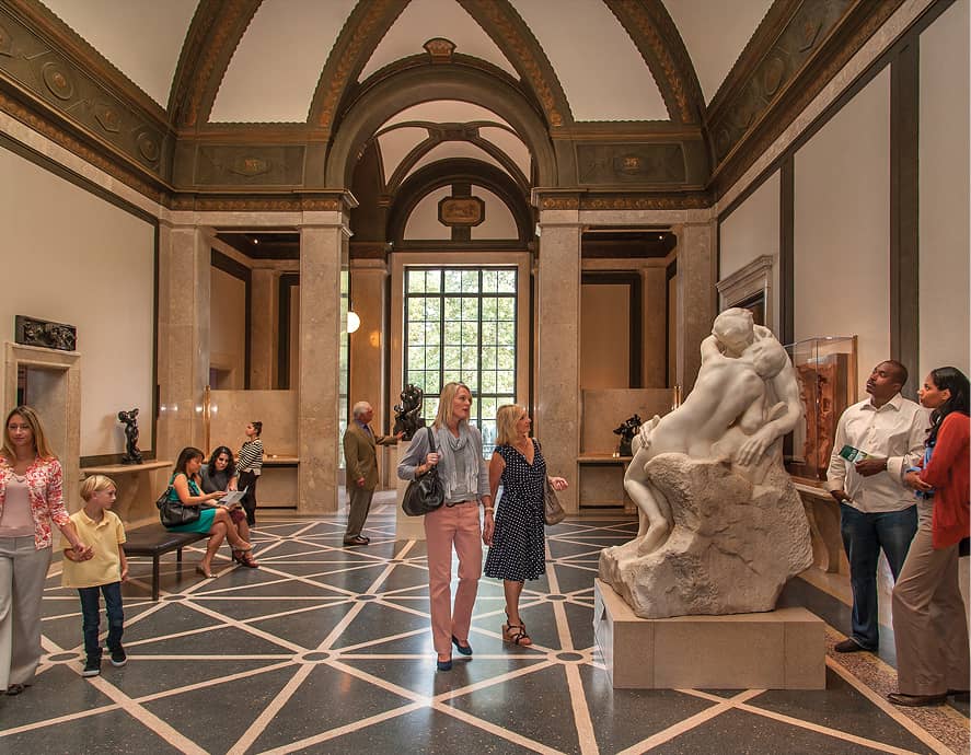 Museum gallery with people observing art. A large marble sculpture of an embracing couple is prominent. The setting is elegant and contemplative.
