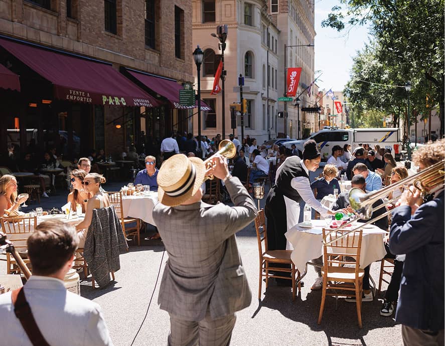 Street scene with diners at outdoor tables enjoying a sunny day. A jazz band plays nearby. The atmosphere is lively and cheerful.