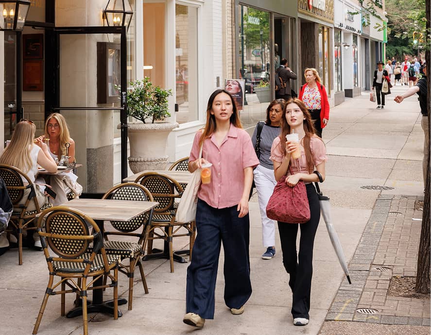 A bustling city sidewalk scene with two young women drinking beverages in the foreground. Tables with seated people line the street, creating a lively atmosphere.