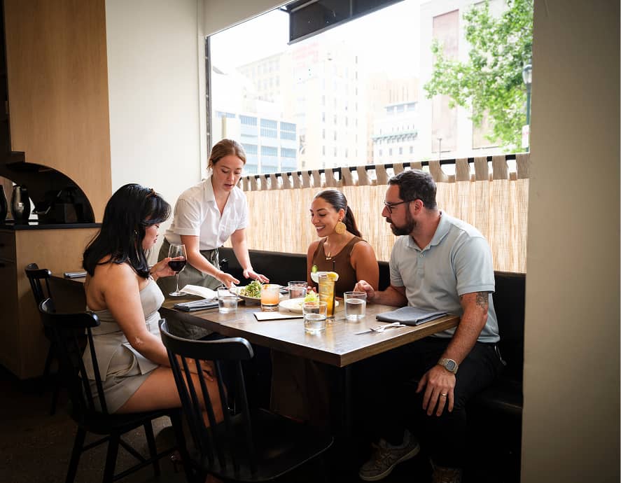 A server in a white shirt delivers a salad to a table where three people are seated, smiling, and chatting. A woman holds a glass of red wine. The setting is a modern, well-lit restaurant with a view of city buildings and trees outside. The atmosphere is casual and friendly.