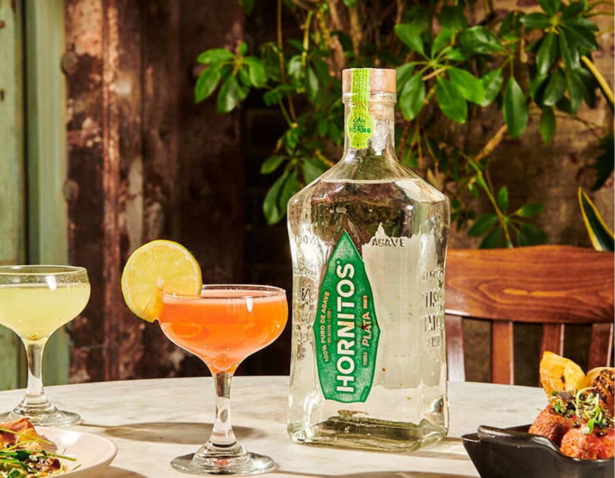 A bottle of Hornitos Plata tequila next to two cocktail glasses, one with an orange drink and lime garnish. The setting is a cozy, warmly-lit dining table with plants in the background.