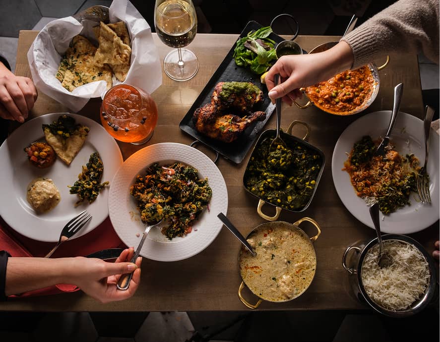 Table with plates of Indian cuisine, including naan, rice, curries, and grilled chicken. Hands serve food, creating a vibrant, communal dining atmosphere.