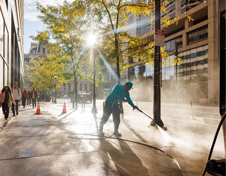 A worker in blue uses a hose to power wash a city sidewalk. Sunlight filters through trees, casting shadows. People walk by, framed by tall buildings.