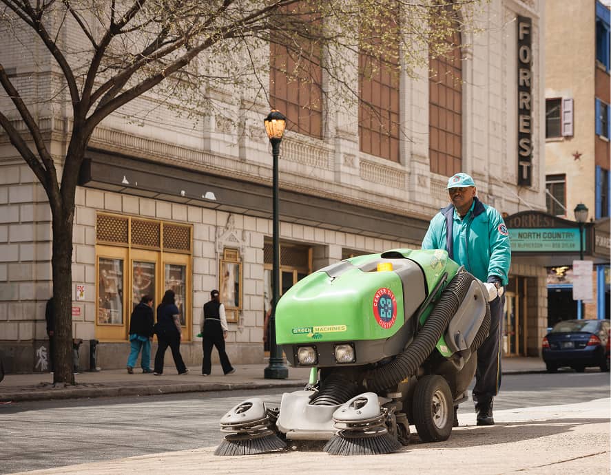 A street cleaner operates a green machine, sweeping a city sidewalk. Nearby buildings and a theater marquee are visible, with pedestrians walking by.