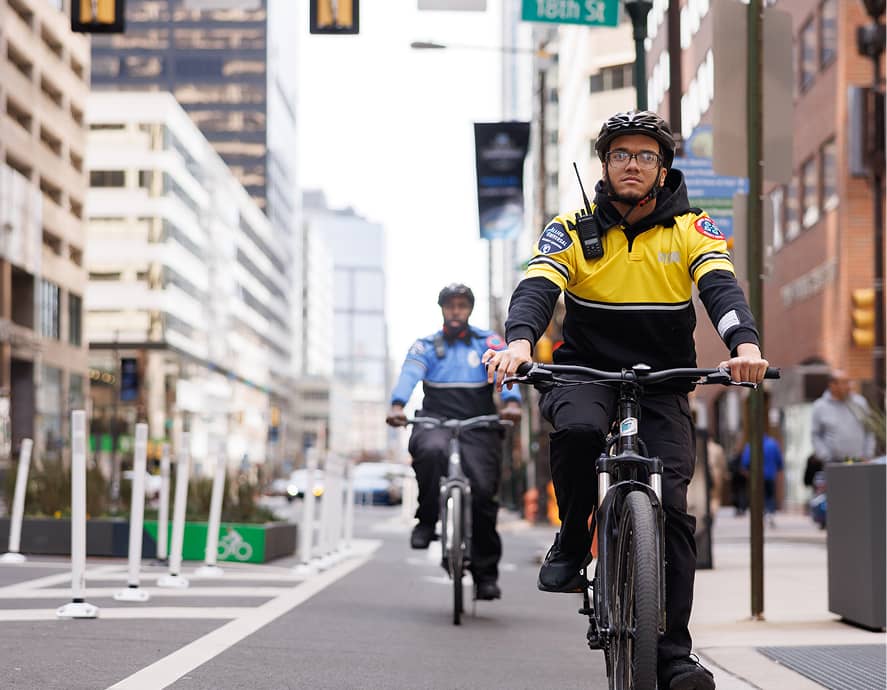 Two uniformed security personnel ride bicycles down a city street. The front cyclist wears a yellow jacket, while the backdrop features tall buildings.