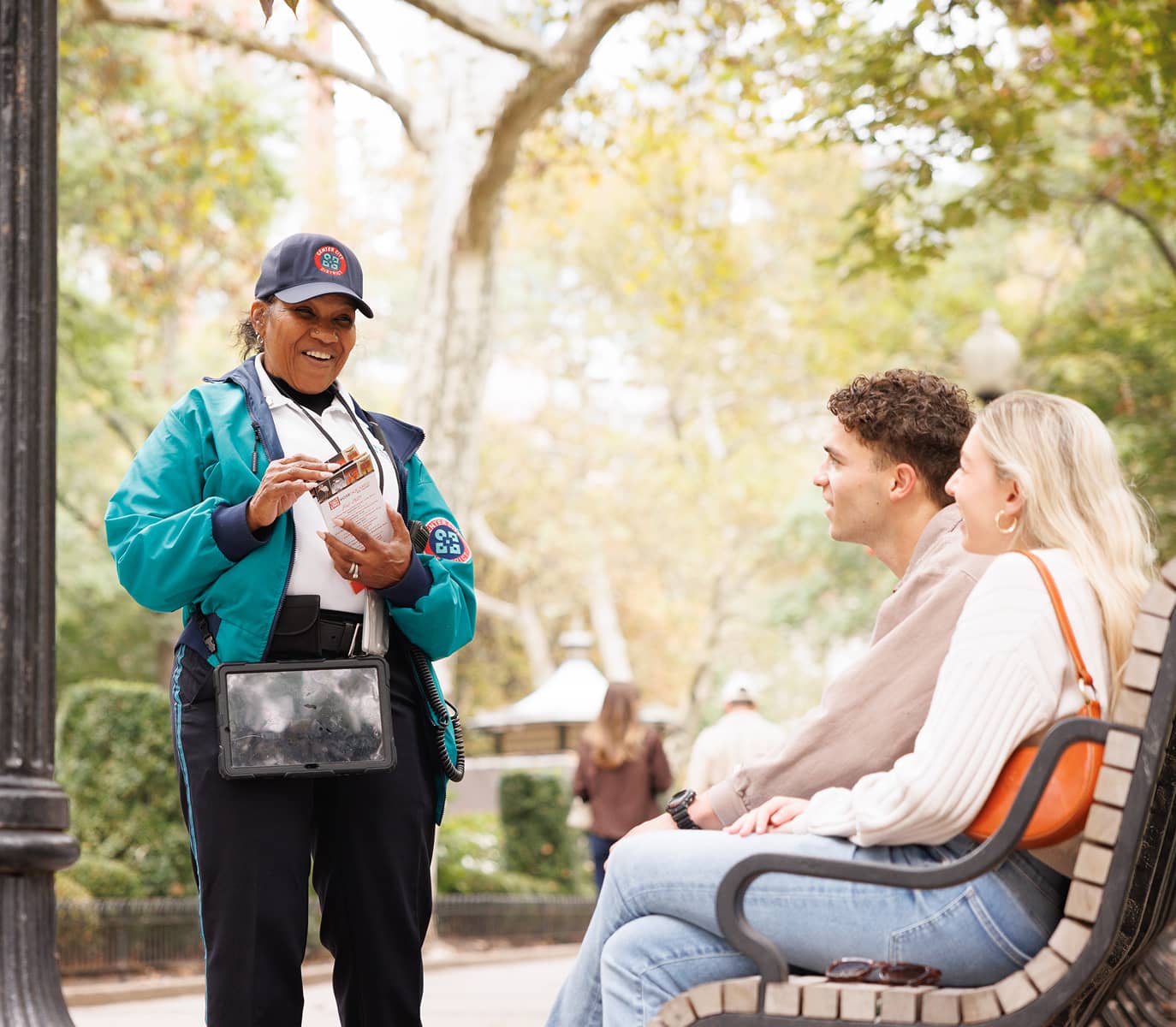 A smiling park employee in a blue uniform engages warmly with a couple seated on a bench, holding maps. A tree-lined park is in the background.