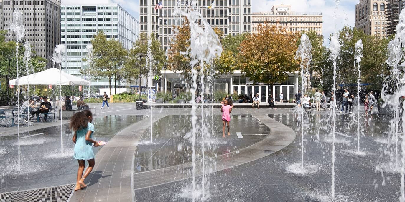 Children play joyfully in a lively urban plaza with water jets, surrounded by tall buildings and trees. The scene conveys a sense of fun and city life.
