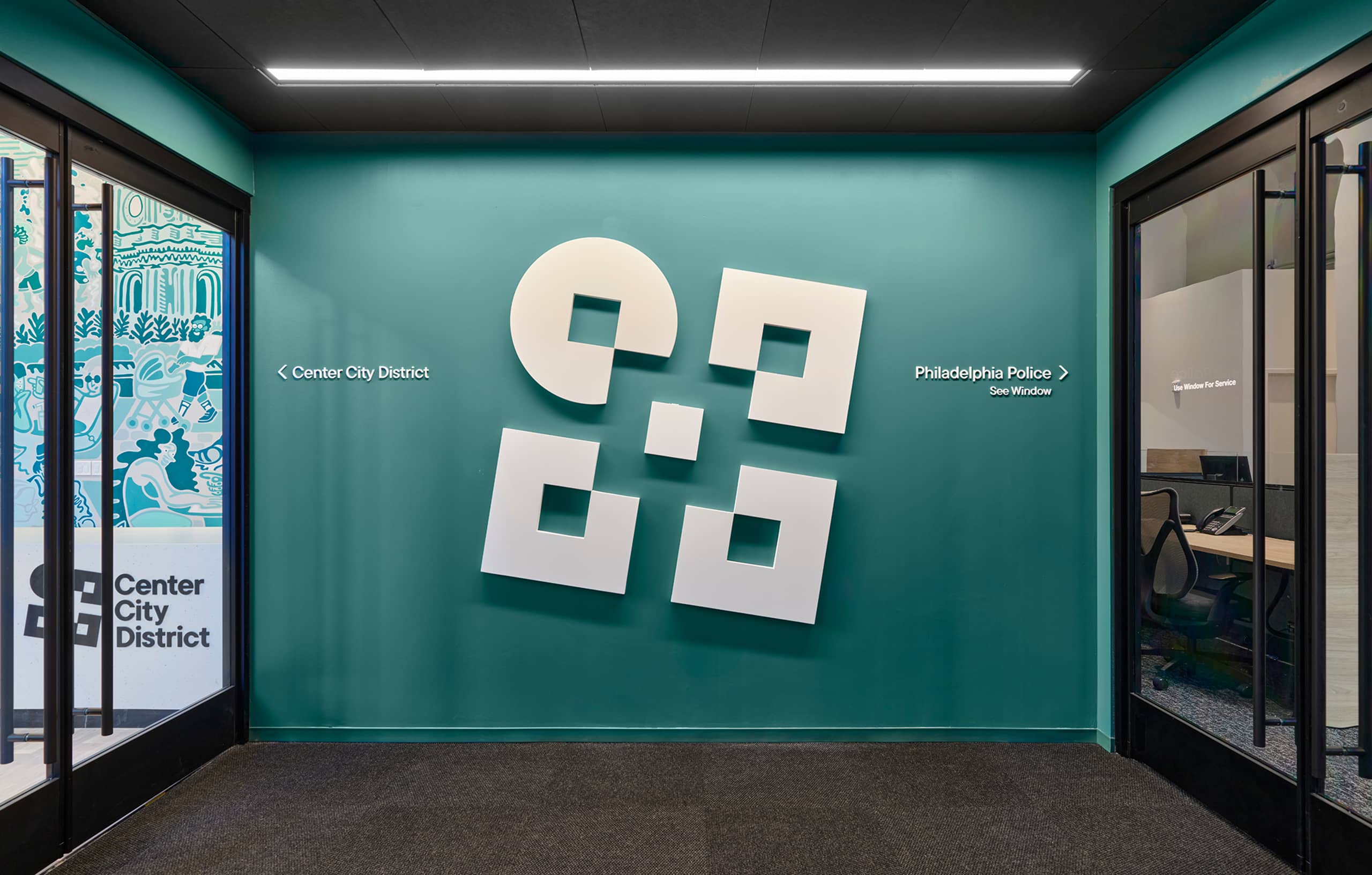 Modern lobby with teal walls featuring a geometric art installation of white squares and circles. Glass doors on each side lead to adjacent offices.