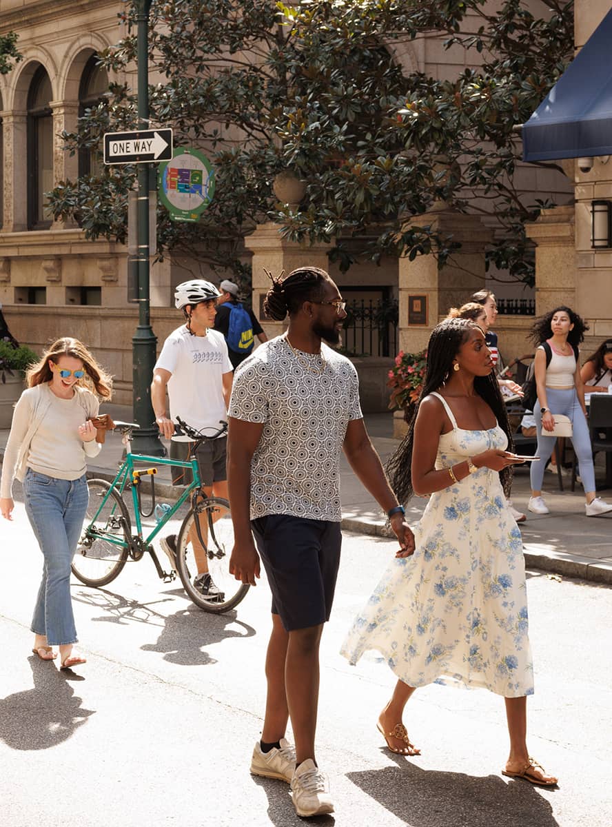 A couple walks hand in hand down a sunny city street, smiling. A cyclist and other pedestrians pass by. Nearby, people enjoy a café, creating a lively scene.