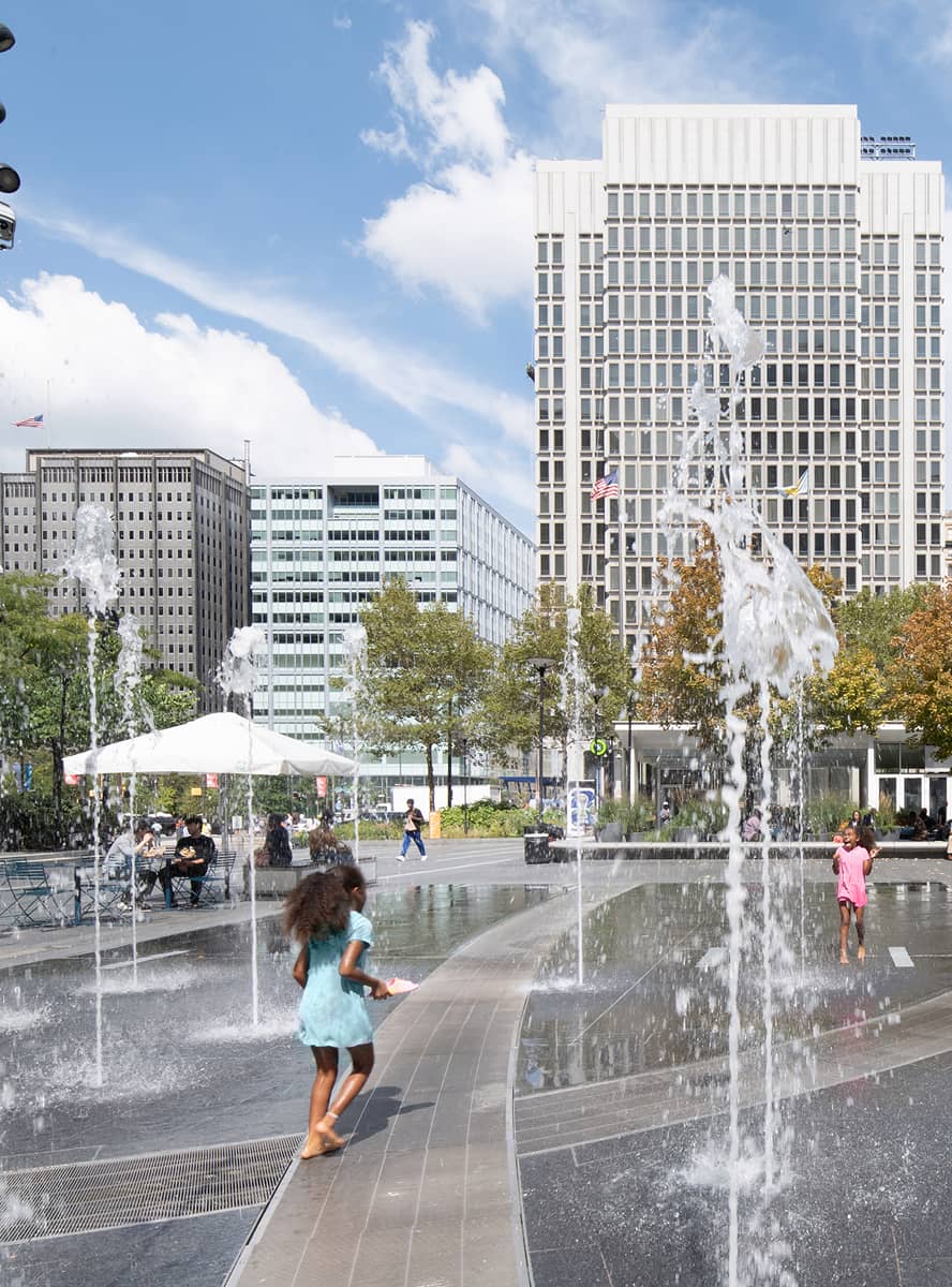 Children play joyfully in a lively urban plaza with water jets, surrounded by tall buildings and trees. The scene conveys a sense of fun and city life.