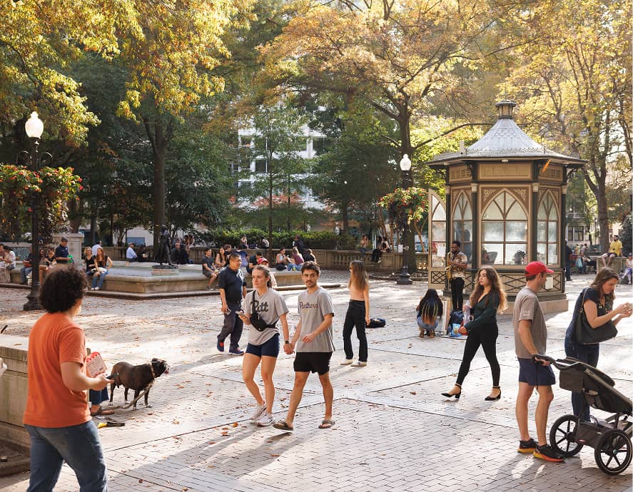 People enjoying a sunny day in a park plaza with a gazebo. Trees with autumn leaves surround them, creating a warm and lively atmosphere.