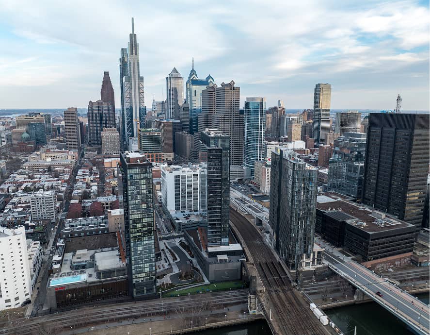 Aerial view of a bustling city skyline with modern skyscrapers under a cloudy sky. Visible roads and railway tracks add to the urban atmosphere.