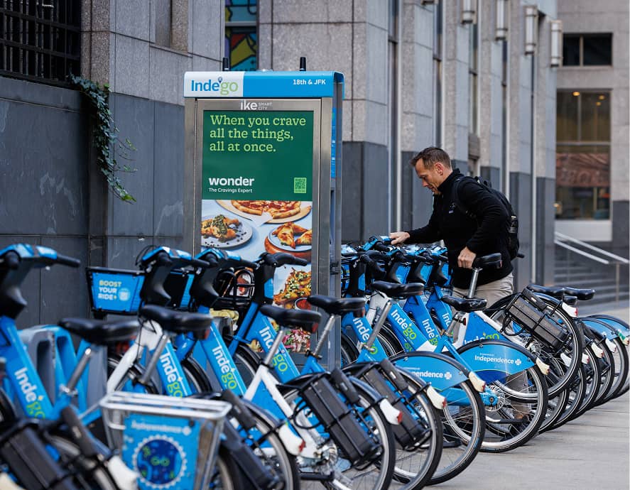A person prepares to rent a blue Indego bike from a bike-sharing station. Nearby, an advert highlights food options, set against an urban street backdrop.