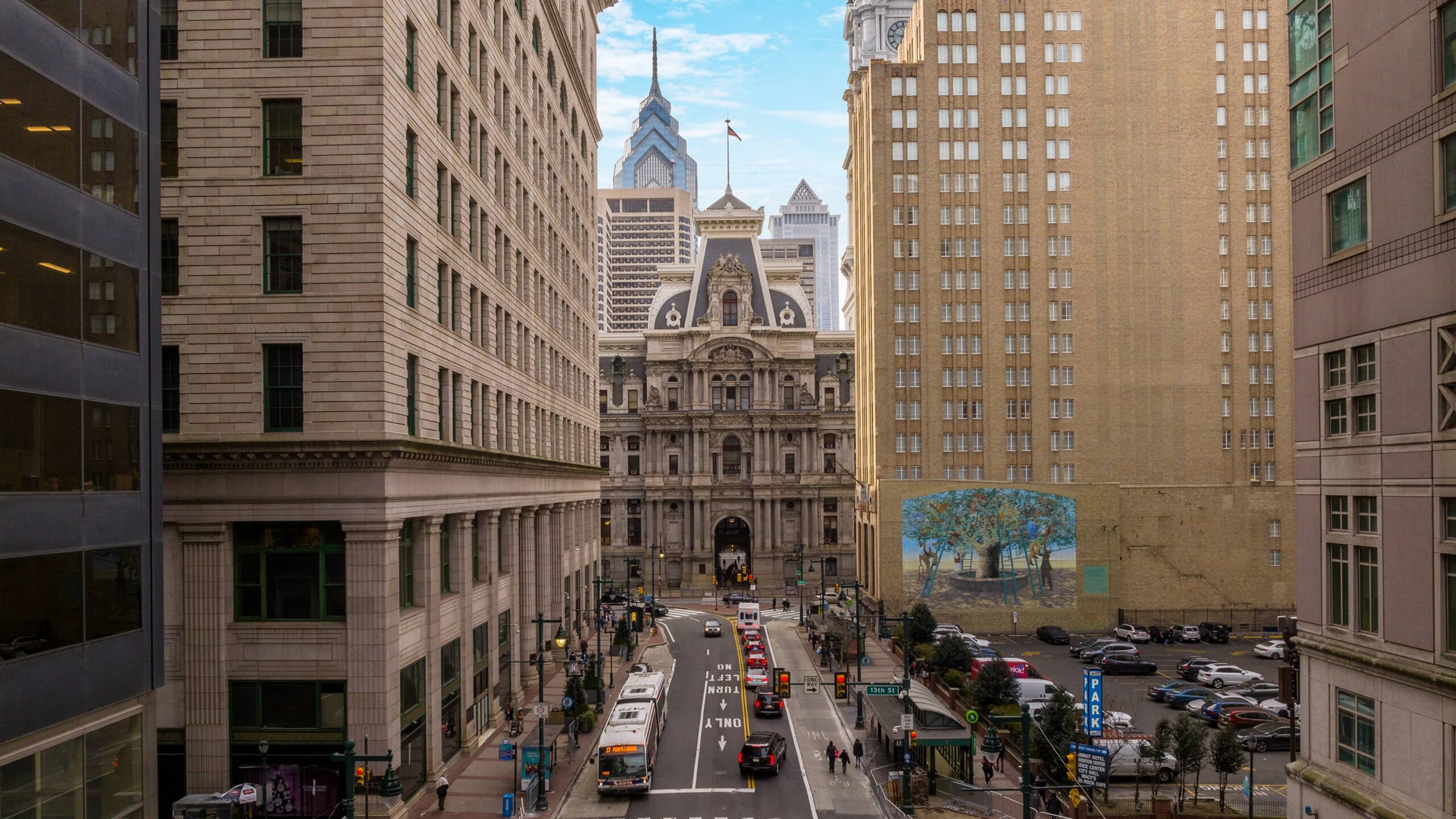 Urban scene with tall buildings flanking a busy street leading to a historic, ornate building. People, cars, and buses create lively city atmosphere.