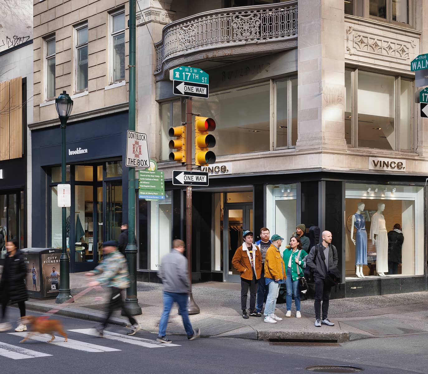 Street scene at a busy corner in front of Vince store. People wait at the crosswalk while a person with a dog walks by. Traffic signs and storefronts visible.