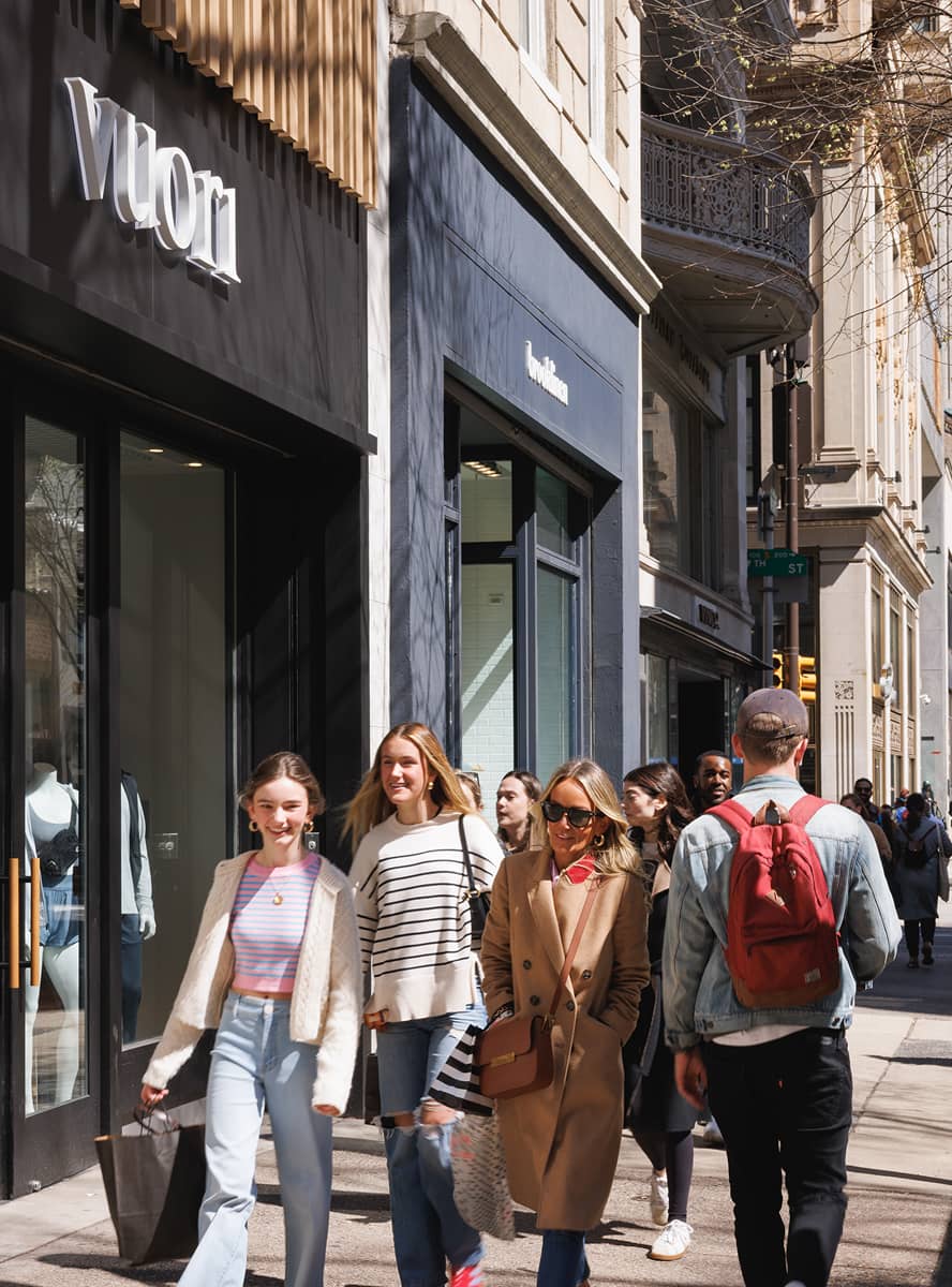A lively city scene shows smiling pedestrians walking past a stylish storefront. The sunny day and casual attire convey a cheerful and relaxed atmosphere.