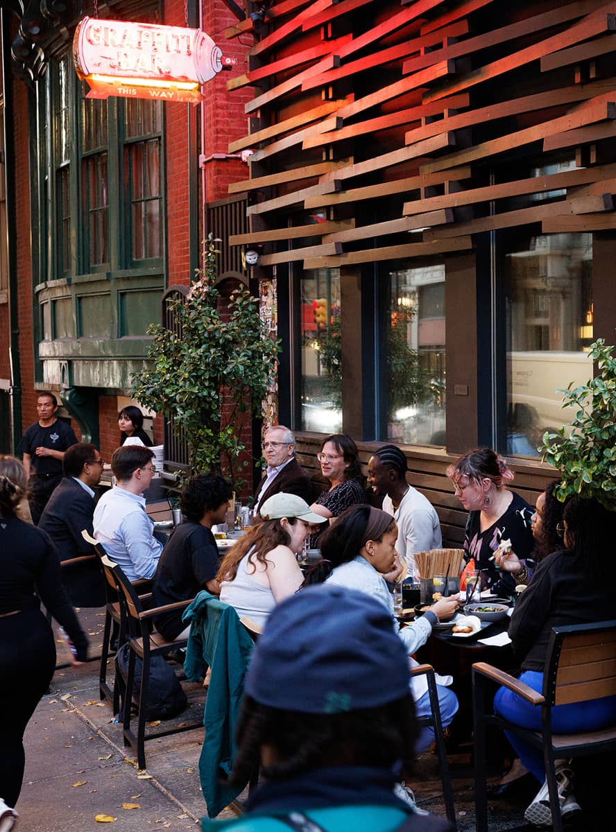 A lively street cafe scene with a group of people dining outdoors. They are engaged in conversation, creating a warm and social atmosphere. Neon sign and plants adorn the setting.