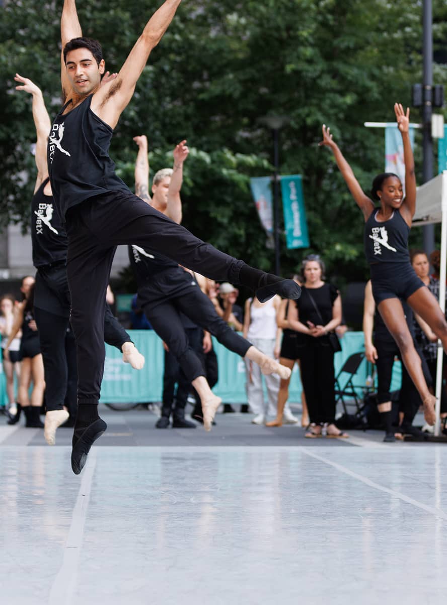 Dancers in black attire perform an energetic jump outdoors. The leading dancer is mid-air, arms extended, creating a dynamic and uplifting atmosphere.