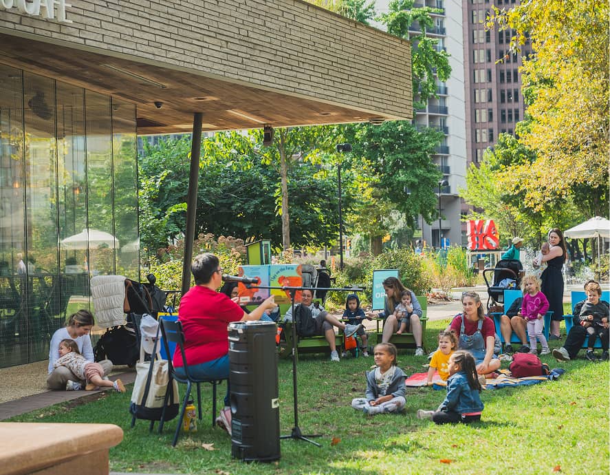 A woman in red leads a storytelling session outdoors. Children and adults sit on the grass listening. The setting is a park next to a glass-walled cafe, with trees and tall buildings in the background, creating a lively and engaging atmosphere.
