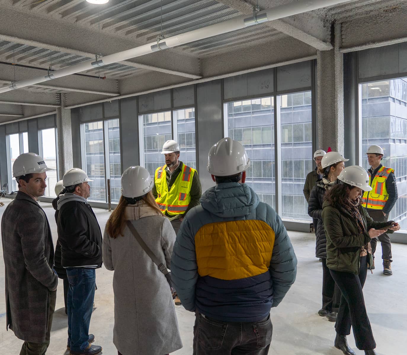 Group of people wearing hard hats and high-visibility vests in a building under construction. They appear engaged in discussion or a tour.