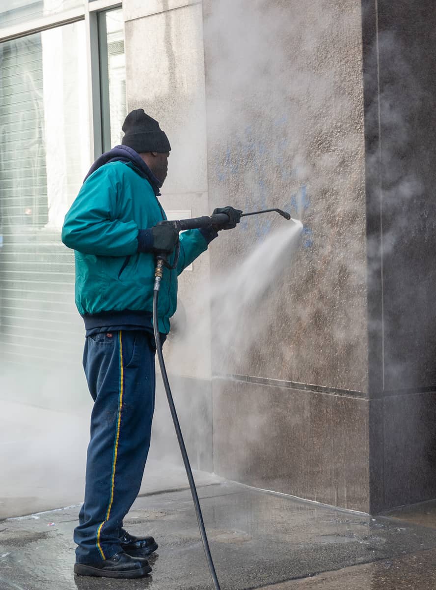 A person in a blue jacket and hat power washes a graffitied wall on a city sidewalk. Dense mist and water spray create a focused, determined mood.