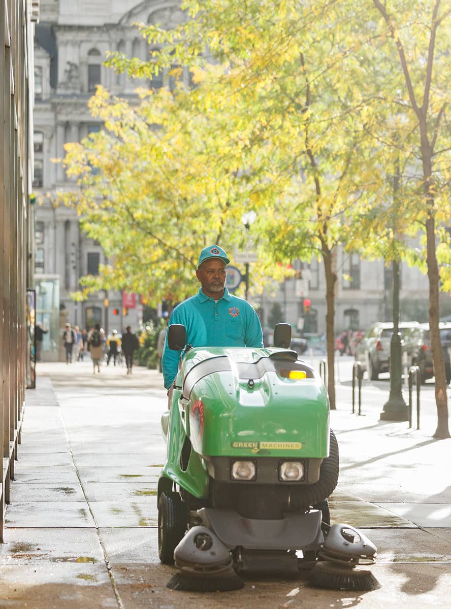 Man in teal uniform operates a green street sweeper on a sunlit city sidewalk. Trees with yellow leaves and historic buildings line the background.