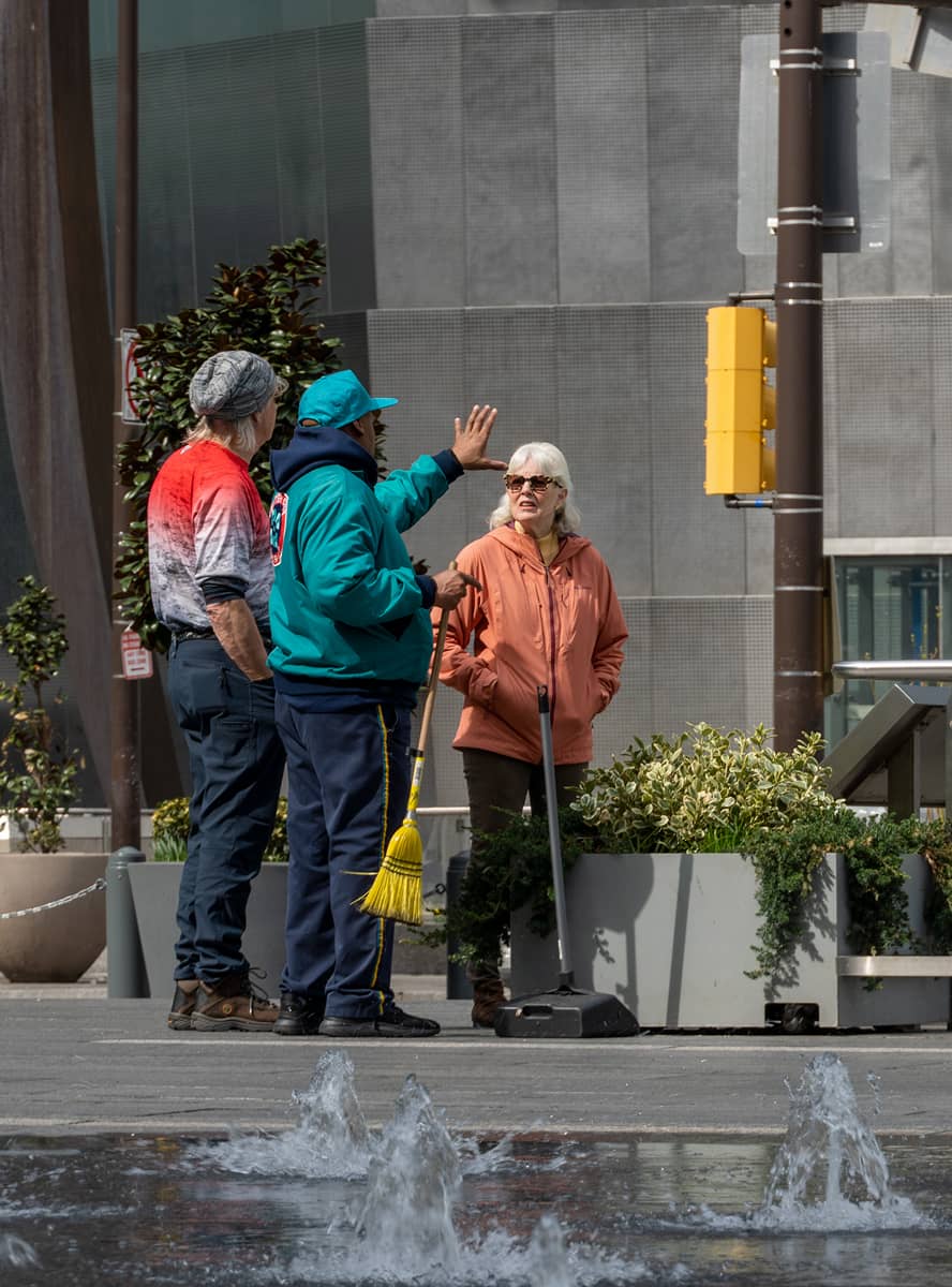 Three people stand on a city street near a fountain. One gestures animatedly while talking. The scene conveys a casual, friendly atmosphere.