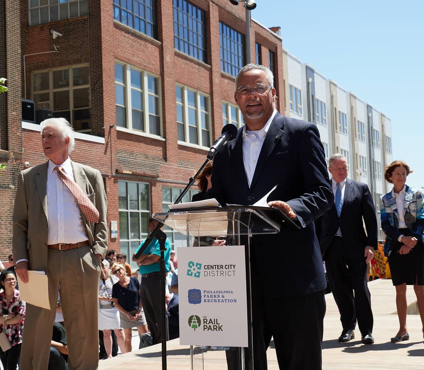 Man speaking at a podium during an outdoor event, with a brick building in the background. Several people stand behind him, some in business attire. Sunny day.