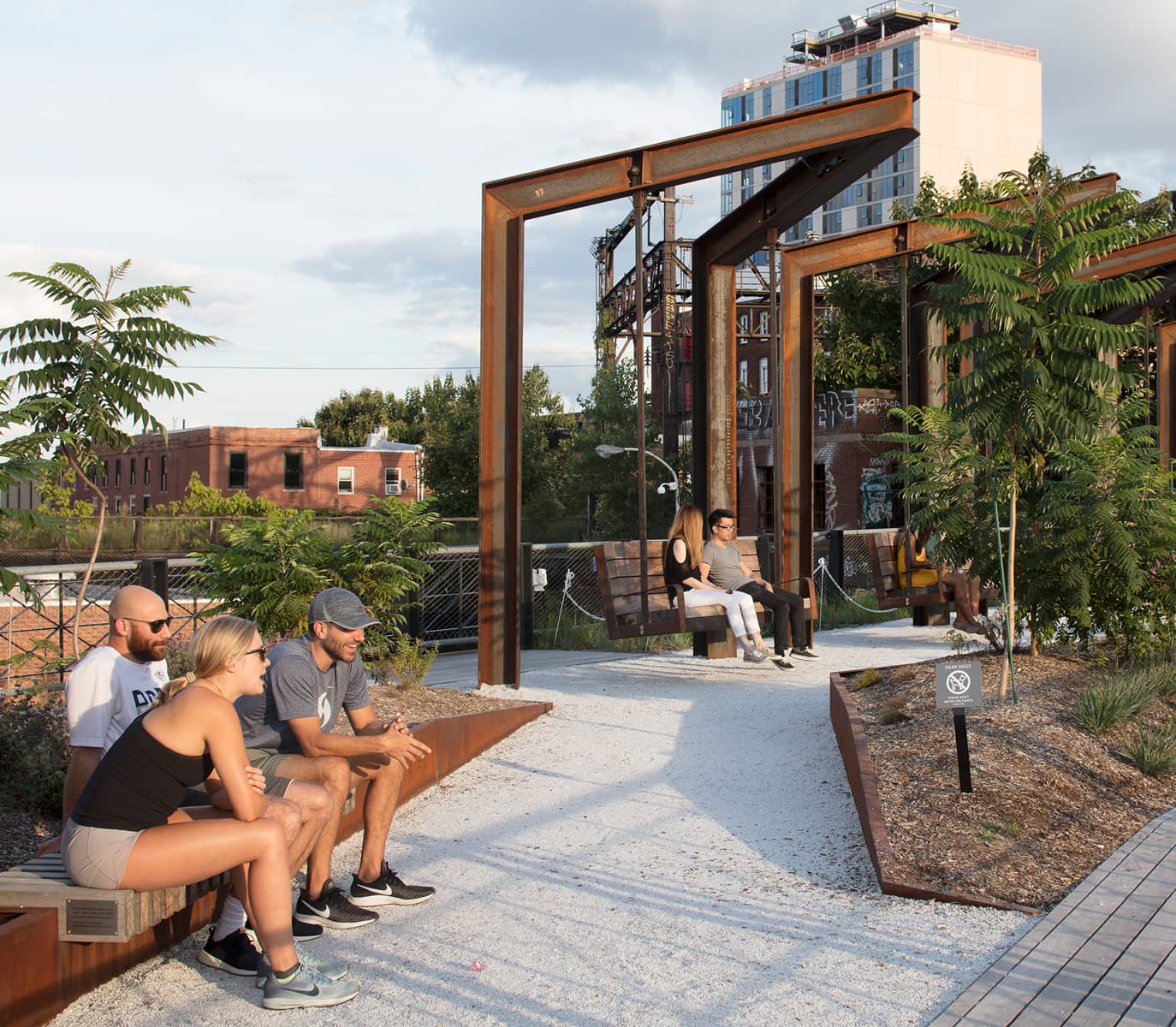 A group of people relax in a green urban park with wooden and metal structures. The atmosphere is calm, with a mix of concrete and foliage.