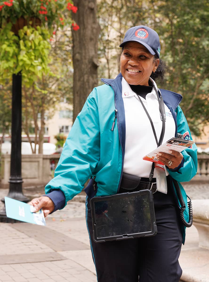 A friendly woman in a teal and white uniform, holding flyers, smiles while handing out information in a park. She wears a cap and carries a tablet.