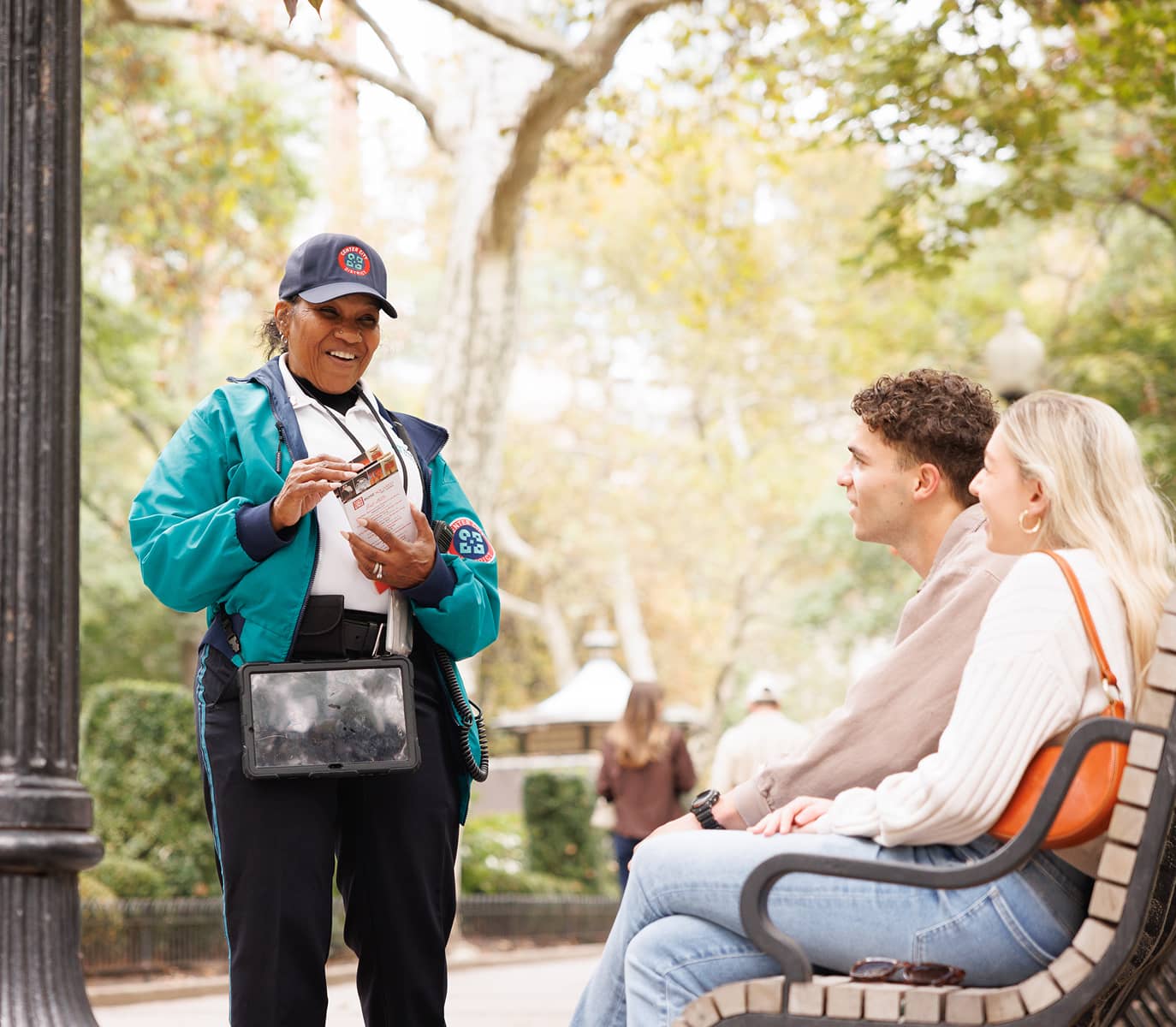 A smiling park employee in a blue uniform engages warmly with a couple seated on a bench, holding maps. A tree-lined park is in the background.