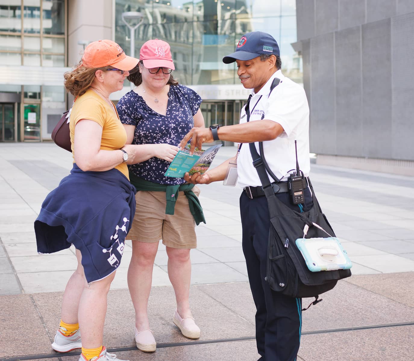 Three people stand outdoors in a city plaza. Two women, wearing casual clothes and hats, consult a map with a smiling security guard dressed in uniform.